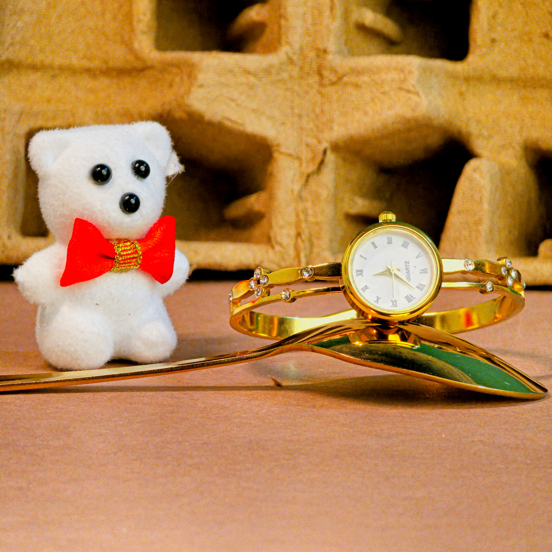 Gold watch with bracelet and small white teddy bear with red bow tie on a textured surface